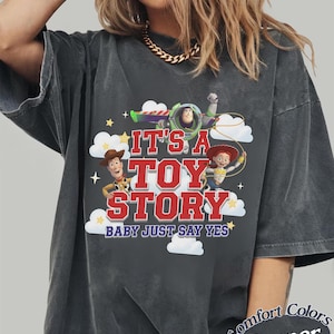 K&ouml;nnte beinhalten: Ein anthrazitfarbenes Kurzarm-T-Shirt mit einem Grafikdruck von Buzz Lightyear, Woody und Jessie aus Toy Story. Der Text lautet IT'S A TOY STORY BABY JUST SAY YES, umgeben von wei&szlig;en Wolken und gelben Sternen auf der Vorderseite des T-Shirts.