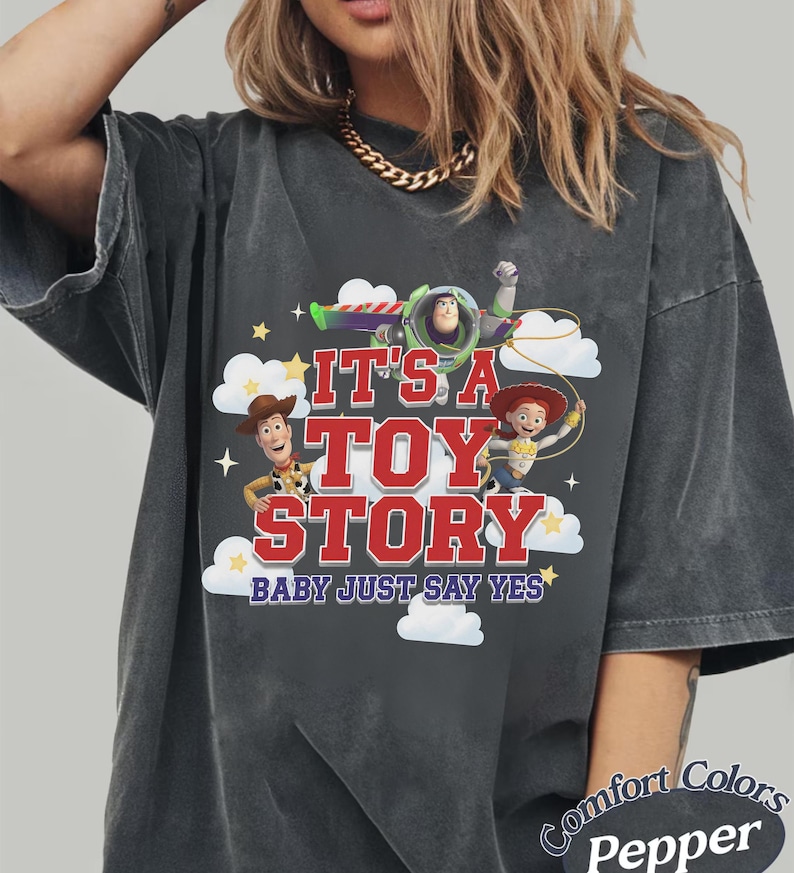 K&ouml;nnte beinhalten: Ein anthrazitfarbenes Kurzarm-T-Shirt mit einem Grafikdruck von Buzz Lightyear, Woody und Jessie aus Toy Story. Der Text lautet IT'S A TOY STORY BABY JUST SAY YES, umgeben von wei&szlig;en Wolken und gelben Sternen auf der Vorderseite des T-Shirts.