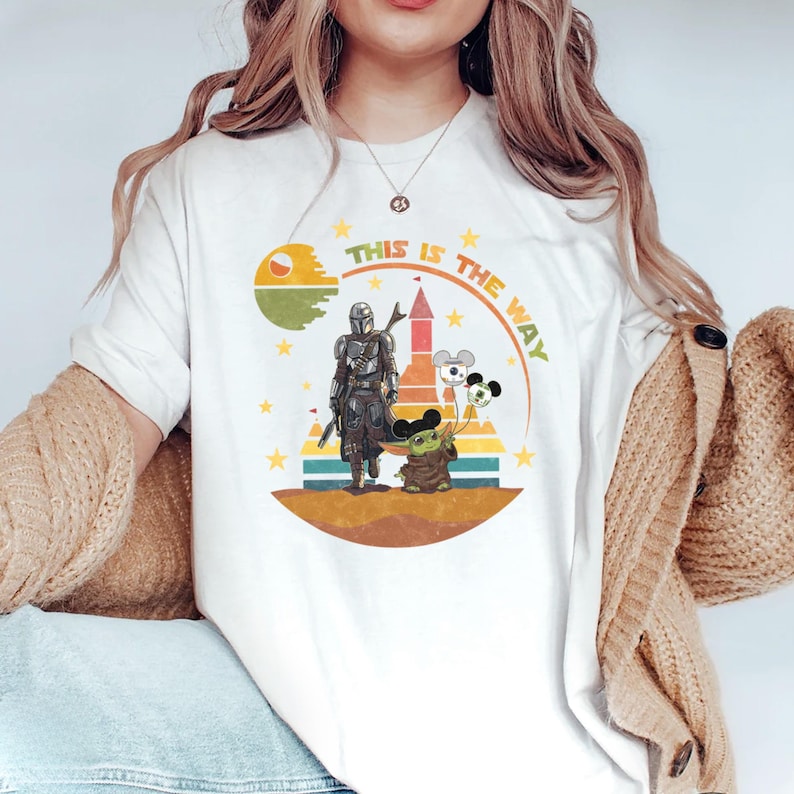 Puede incluir: Camiseta blanca con un dise&ntilde;o gr&aacute;fico con el texto "THIS IS THE WAY". El dise&ntilde;o incluye una figura mandaloriana, un personaje de Grogu y globos con forma de Mickey Mouse. El dise&ntilde;o est&aacute; ambientado sobre un fondo de un planeta y estrellas.