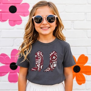 K&ouml;nnte beinhalten: Ein anthrazitfarbenes Kurzarm-T-Shirt mit einem Grafikdruck von zwei kastanienbraunen Cowboystiefeln mit wei&szlig;en Wirbelmustern. Die Stiefel sind nebeneinander in der Mitte des T-Shirts platziert und zeigen detaillierte N&auml;hte.