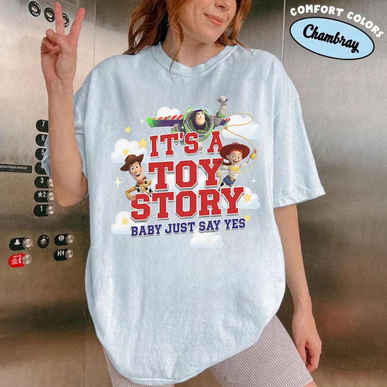 K&ouml;nnte beinhalten: Ein hellblaues Kurzarm-T-Shirt mit einem Grafikdruck von Woody, Buzz Lightyear und Jessie aus Toy Story. Der Text lautet IT'S A TOY STORY BABY JUST SAY YES. Das Design enth&auml;lt wei&szlig;e Wolken und gelbe Sterne auf einem chambrayfarbenen Stoff.