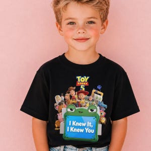 May include: A black short-sleeved t-shirt featuring a colorful graphic print of Toy Story 5 characters. The design includes a green frog-shaped frame containing the text "I Knew It, I Knew You" in white letters against a blue background.
