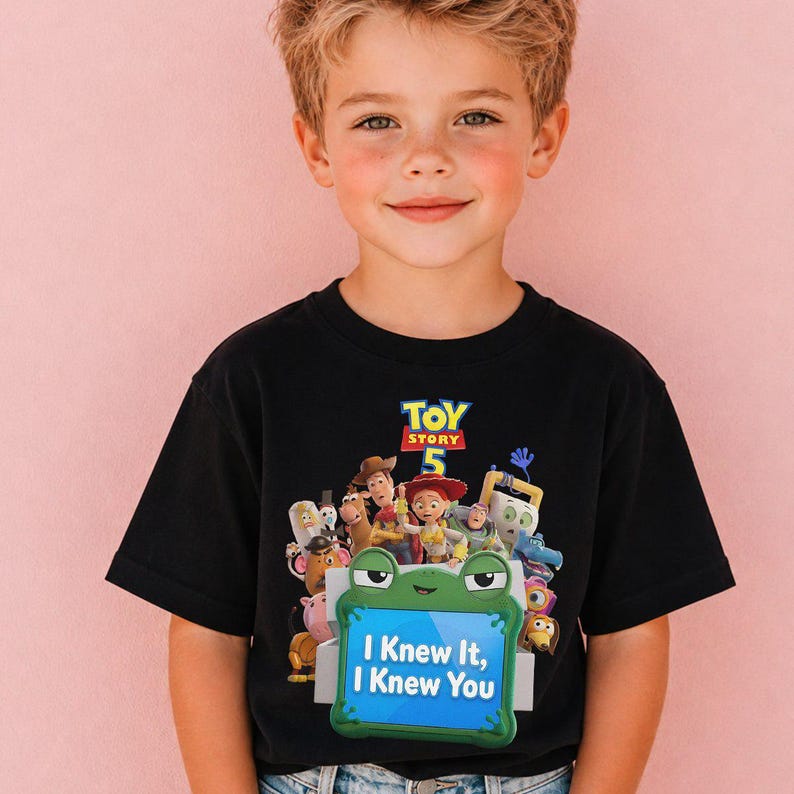 May include: A black short-sleeved t-shirt featuring a colorful graphic print of Toy Story 5 characters. The design includes a green frog-shaped frame containing the text "I Knew It, I Knew You" in white letters against a blue background.
