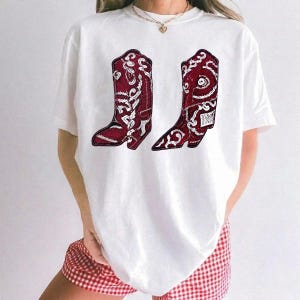 K&ouml;nnte beinhalten: Ein wei&szlig;es kurz&auml;rmeliges T-Shirt mit einem Grafikdruck von zwei dunkelroten Cowboystiefeln mit komplizierten wei&szlig;en Blumen- und Paisley-Mustern. Das T-Shirt hat einen Rundhalsausschnitt und wird mit einer rot-wei&szlig; karierten Shorts kombiniert.