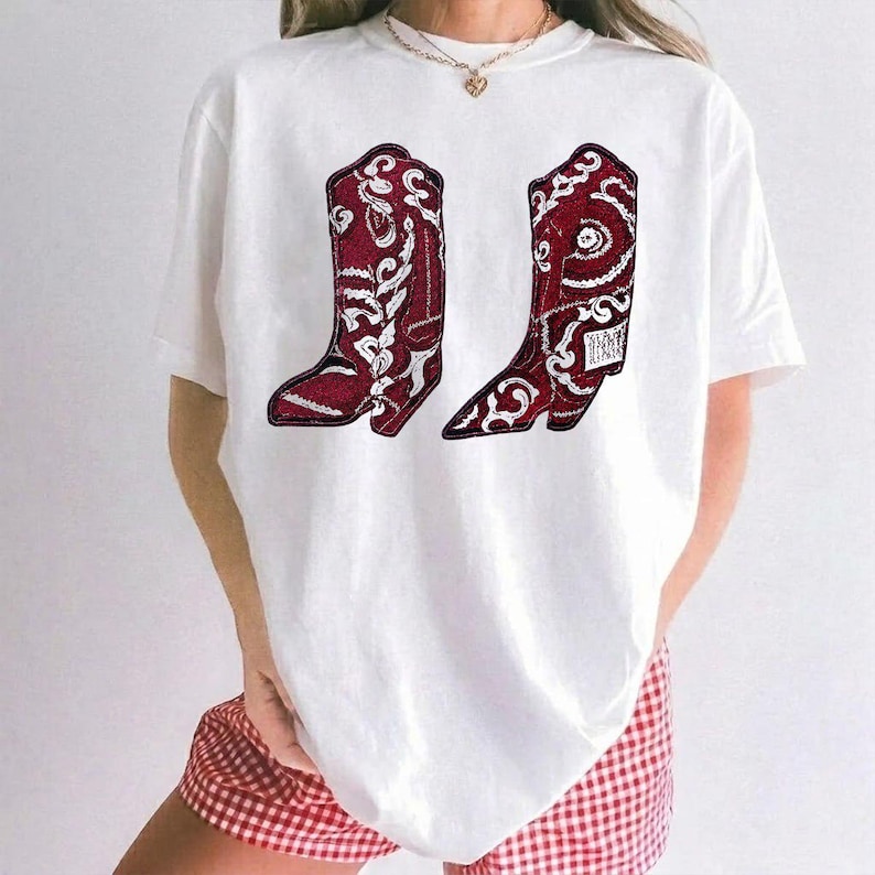 K&ouml;nnte beinhalten: Ein wei&szlig;es kurz&auml;rmeliges T-Shirt mit einem Grafikdruck von zwei dunkelroten Cowboystiefeln mit komplizierten wei&szlig;en Blumen- und Paisley-Mustern. Das T-Shirt hat einen Rundhalsausschnitt und wird mit einer rot-wei&szlig; karierten Shorts kombiniert.