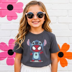 May include: A charcoal gray short-sleeve t-shirt featuring a graphic print of a blue alien character wearing a red and blue spider-themed costume. The character has large white eyes, a spider emblem on the chest, and is making a hand gesture.