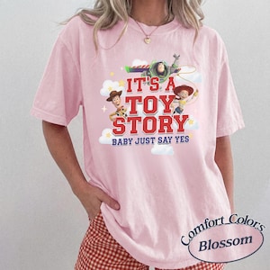 K&ouml;nnte beinhalten: Ein hellrosa T-Shirt mit kurzen &Auml;rmeln und einem Grafikdruck von Buzz Lightyear, Woody und Jessie aus Toy Story. Der Text lautet IT'S A TOY STORY BABY JUST SAY YES, erg&auml;nzt durch wei&szlig;e Wolken und gelbe Sterne auf weichem Baumwollstoff.