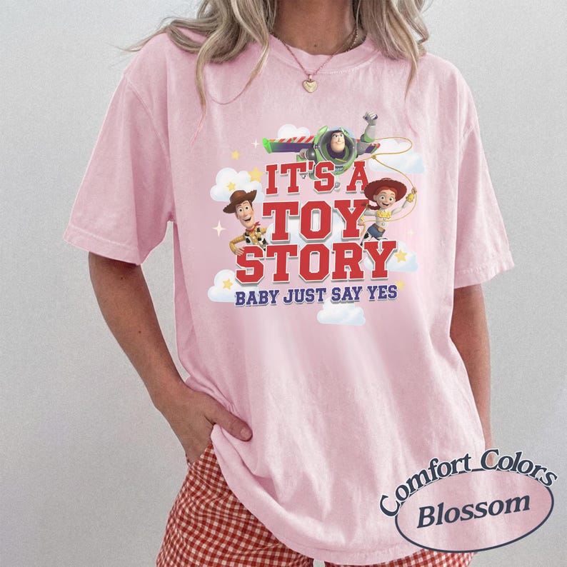 K&ouml;nnte beinhalten: Ein hellrosa T-Shirt mit kurzen &Auml;rmeln und einem Grafikdruck von Buzz Lightyear, Woody und Jessie aus Toy Story. Der Text lautet IT'S A TOY STORY BABY JUST SAY YES, erg&auml;nzt durch wei&szlig;e Wolken und gelbe Sterne auf weichem Baumwollstoff.