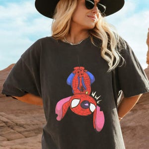May include: A charcoal gray short-sleeve t-shirt featuring a graphic print of a character resembling Stitch wearing a red and blue spider-themed costume. The character is depicted hanging upside down with large ears and white eyes.