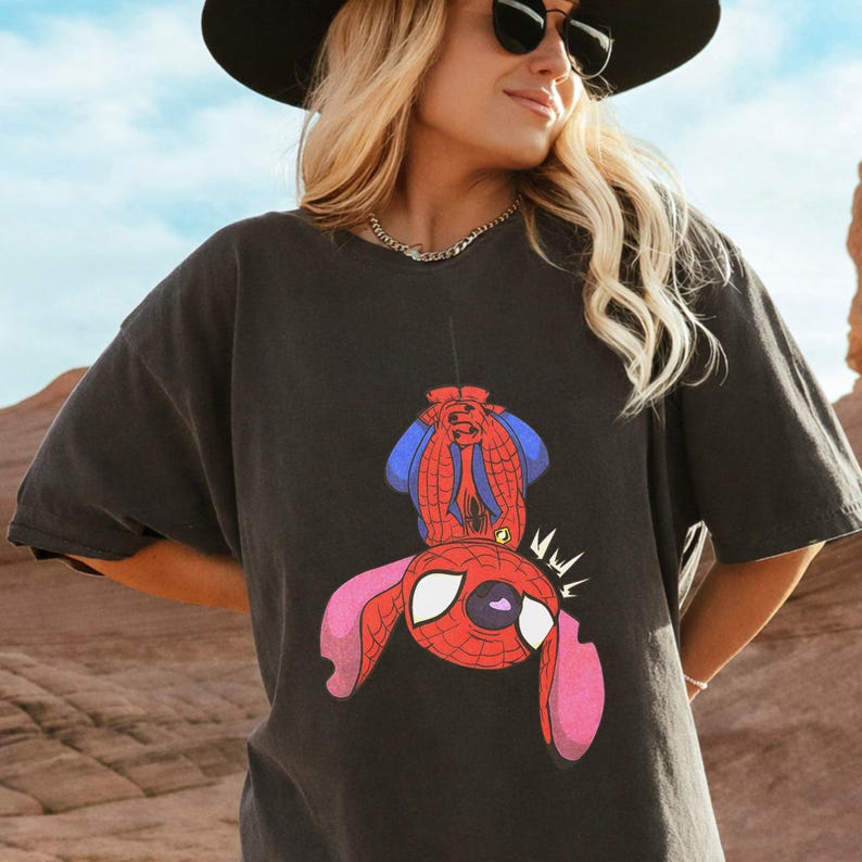 May include: A charcoal gray short-sleeve t-shirt featuring a graphic print of a character resembling Stitch wearing a red and blue spider-themed costume. The character is depicted hanging upside down with large ears and white eyes.