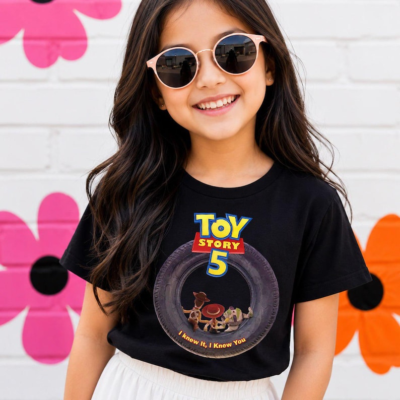 May include: A black short-sleeved t-shirt featuring a graphic of a tire with the text Toy Story 5 and the phrase I knew it, I knew you. Inside the tire graphic are small figures of Woody, Jessie, and Buzz Lightyear from the Toy Story franchise.