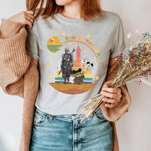 Puede incluir: Camiseta gris con un dise&ntilde;o de The Mandalorian y Grogu, con el texto "THIS IS THE WAY". El dise&ntilde;o incluye un planeta, un castillo y globos de Mickey Mouse. La camiseta es de cuello redondo.