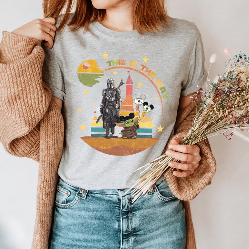 Puede incluir: Camiseta gris con un dise&ntilde;o de The Mandalorian y Grogu, con el texto "THIS IS THE WAY". El dise&ntilde;o incluye un planeta, un castillo y globos de Mickey Mouse. La camiseta es de cuello redondo.