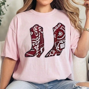 K&ouml;nnte beinhalten: Ein hellrosa T-Shirt mit kurzen &Auml;rmeln und einem Grafikdruck von zwei Cowboystiefeln. Die Stiefel sind dunkelrot mit komplizierten wei&szlig;en Paisley- und Blumenmustern und haben eine strukturierte Optik, die an Pailletten oder Stickereien erinnert.