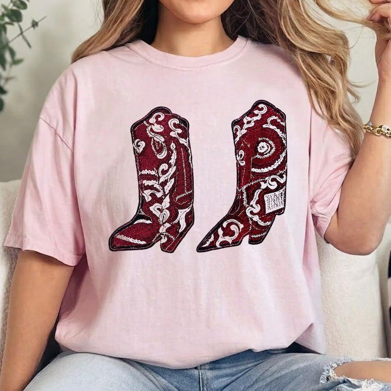 K&ouml;nnte beinhalten: Ein hellrosa T-Shirt mit kurzen &Auml;rmeln und einem Grafikdruck von zwei Cowboystiefeln. Die Stiefel sind dunkelrot mit komplizierten wei&szlig;en Paisley- und Blumenmustern und haben eine strukturierte Optik, die an Pailletten oder Stickereien erinnert.