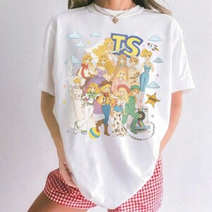 May include: A white short-sleeve t-shirt featuring a colorful graphic print of various animated characters in a retro style. The design includes the letters TS at the top, a yellow star, clouds, and a rocket ship, all arranged in a group illustration.