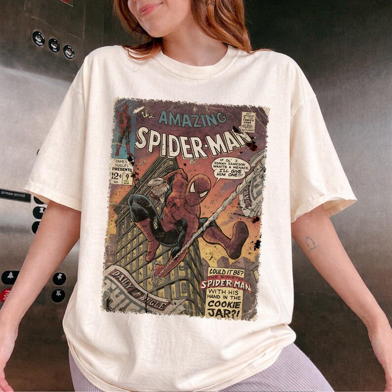 May include: Cream-colored t-shirt featuring a vintage comic book cover of Spider-Man. The graphic shows Spider-Man swinging through a city, with text that reads "Amazing Spider-Man" and "Daily Bugle".