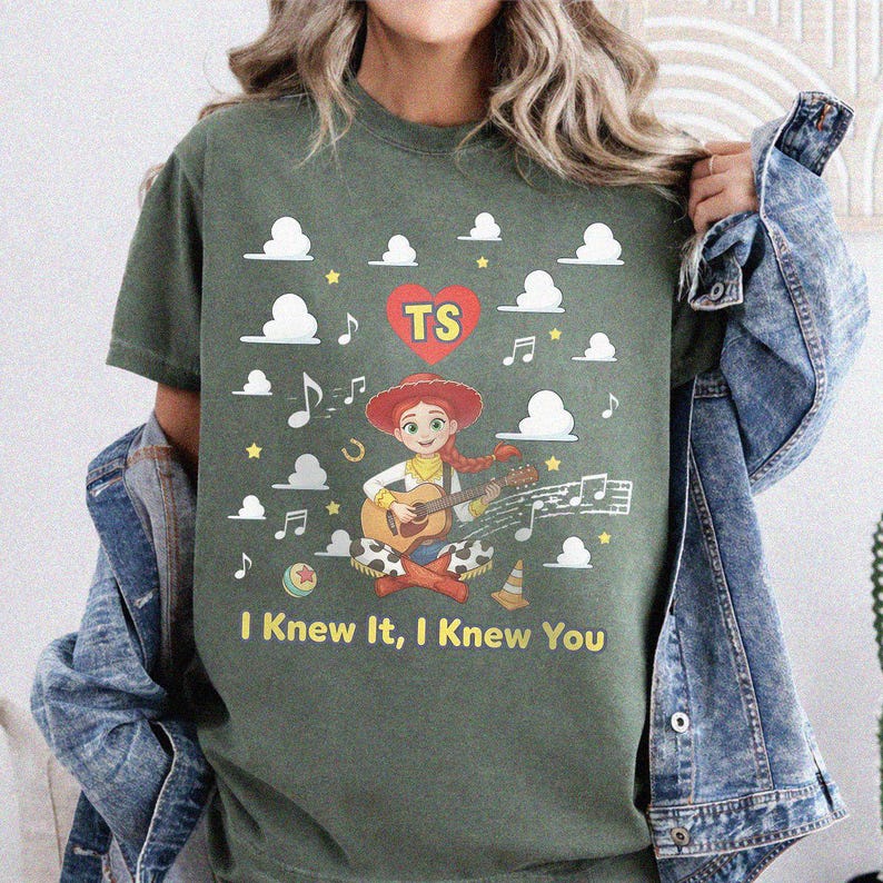 May include: An olive green t-shirt featuring a graphic of a cartoon cowgirl playing an acoustic guitar. The design includes white clouds, musical notes, a red heart with the letters TS, and the yellow text I Knew It, I Knew You.