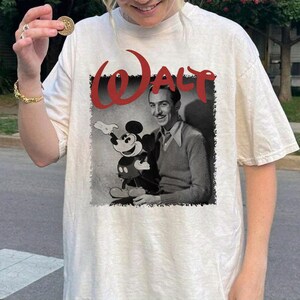 May include: Cream-coloured t-shirt featuring a black and white image of Walt Disney with Mickey Mouse. The word "WALT" is printed in red above the image. The shirt has short sleeves and a relaxed fit.