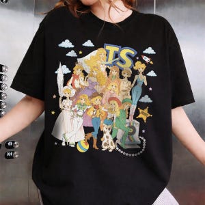 May include: A black short-sleeve t-shirt featuring a graphic print of multiple blonde characters in various outfits. The design includes the letters TS, a rocket, clouds, a star labeled SHERIFF, a ball, and a camera. The characters are depicted in a cartoon style with diverse clothing items like hats, dresses, and pants.