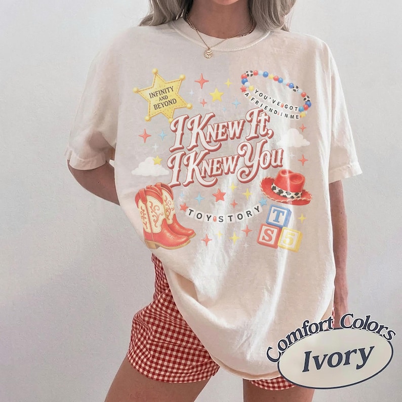 May include: An ivory short-sleeve t-shirt featuring a graphic print with the text "I Knew It, I Knew You", "Infinity and Beyond", "Toy Story", and "You've Got a Friend in Me". The design includes a yellow star, cowboy boots, a hat, and blocks.
