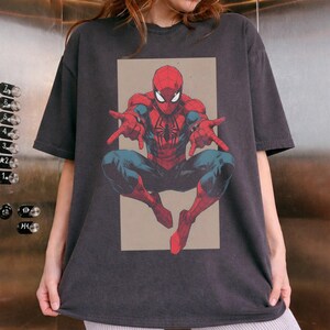 Pu&ograve; includere: Una t-shirt a maniche corte color grigio antracite con un'illustrazione grafica centrata del personaggio dei fumetti Spider-Man in posizione accovacciata. Il personaggio indossa un costume rosso e blu con un emblema a ragno nero sul petto.
