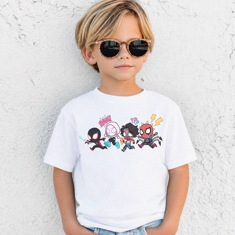 May include: A white short-sleeved t-shirt featuring a colorful graphic print of four chibi-style superhero characters running. The design includes comic book elements like the text BAM, question marks, and a lightning bolt in bright colors.