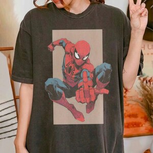 Puede incluir: Una camiseta de manga corta gris carb&oacute;n con un estampado gr&aacute;fico del personaje de c&oacute;mic Spider-Man en posici&oacute;n agachada. El personaje viste un traje rojo y azul con un emblema de ara&ntilde;a negra en el pecho, sobre un fondo rectangular de color beige.