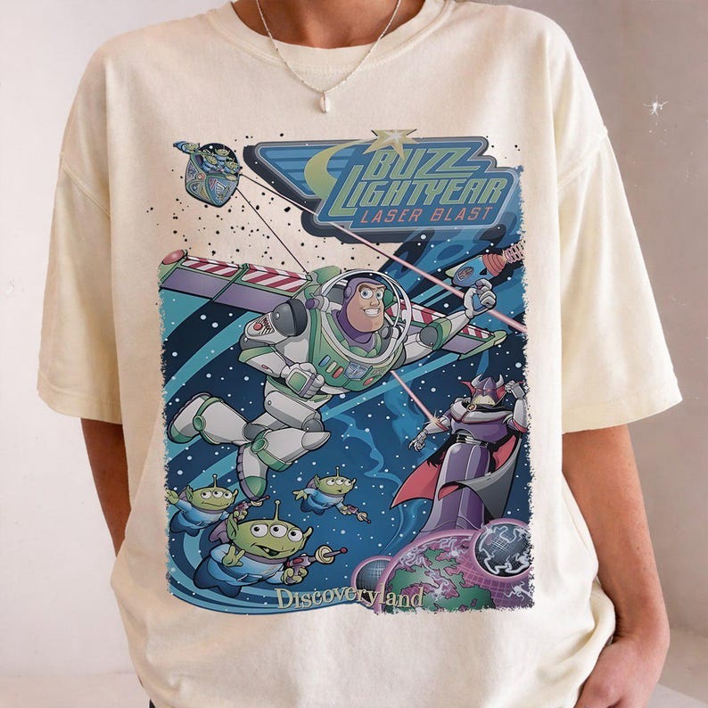 May include: Cream-colored t-shirt featuring a colorful graphic of Buzz Lightyear in a space scene with the text "Buzz Lightyear Laser Blast" and "Discoveryland". The design includes aliens and Zurg.