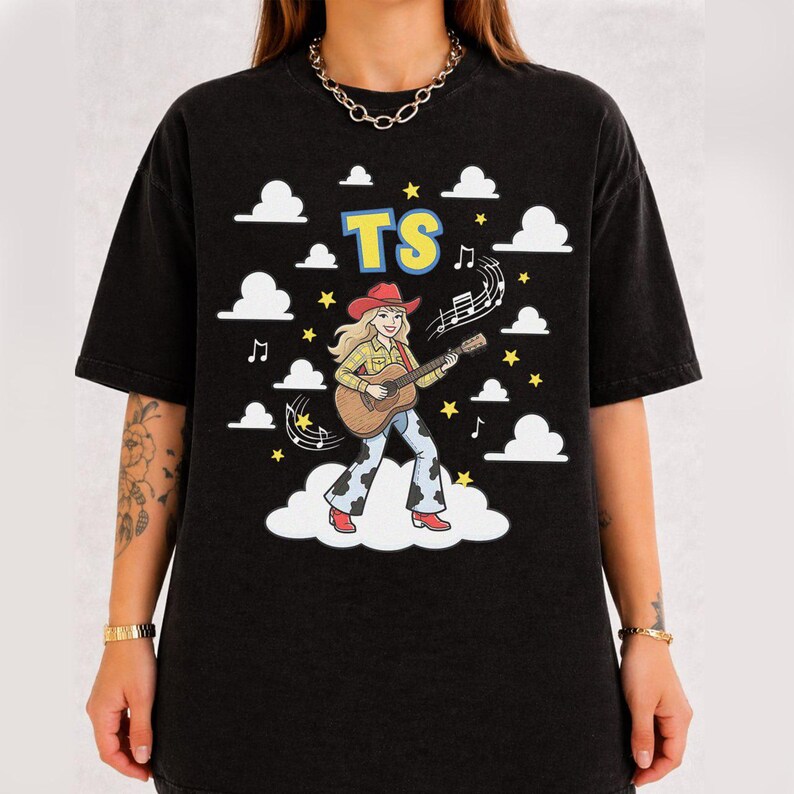 May include: A black short-sleeve t-shirt featuring a graphic of a blonde person in a red cowboy hat, yellow plaid shirt, and cow-print trousers playing an acoustic guitar. The design includes white clouds, yellow stars, musical notes, and the text TS.