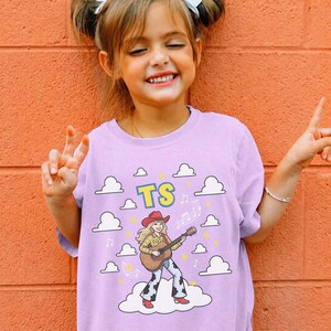 May include: A light purple t-shirt featuring a graphic of a cartoon character wearing a red cowboy hat, a yellow plaid shirt, and cow-print trousers while playing an acoustic guitar. The design includes white clouds, yellow stars, and the letters TS.