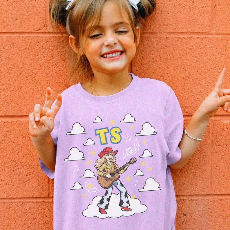 May include: A light purple t-shirt featuring a graphic of a cartoon character wearing a red cowboy hat, a yellow plaid shirt, and cow-print trousers while playing an acoustic guitar. The design includes white clouds, yellow stars, and the letters TS.