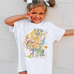 May include: A white short-sleeve t-shirt featuring a colorful graphic print of multiple illustrated characters with blonde hair, blue clouds, stars, and the letters TS in a bold blue font. The design includes various outfits and accessories.