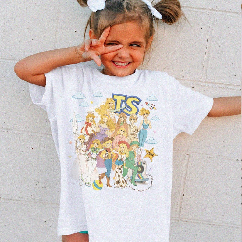 May include: A white short-sleeve t-shirt featuring a colorful graphic print of multiple illustrated characters with blonde hair, blue clouds, stars, and the letters TS in a bold blue font. The design includes various outfits and accessories.