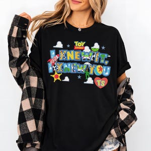 Pode incluir: Uma t-shirt preta com um gr&aacute;fico colorido de Toy Story e o texto I KNEW IT, I KNEW YOU. O design inclui um foguete, uma estrela de xerife, um soldado de paraquedas e um cora&ccedil;&atilde;o vermelho com as letras TS, sobre uma camisa de flanela xadrez.