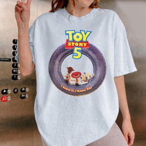 May include: A light blue t-shirt featuring a graphic of a black tire with the text Toy Story 5 in yellow and red. Inside the tire are illustrations of Woody, Jessie, and Buzz Lightyear, with the text I knew it, I knew you printed below.