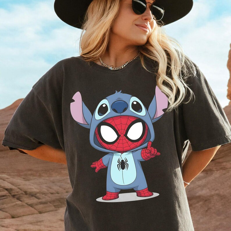 May include: A charcoal gray short-sleeve t-shirt featuring a graphic print of a blue alien character wearing a red and blue spider-themed costume with a spider emblem on the chest and a hood pulled up over its head, displaying large white eyes.