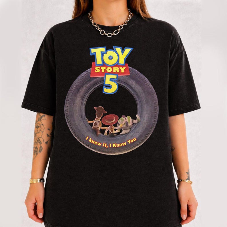 May include: A black short-sleeve t-shirt featuring a graphic of a large tire with the text Toy Story 5 above it. Inside the tire are illustrations of Woody, Jessie, and Buzz Lightyear, with the text I knew it, I knew You printed below them.