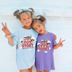 K&ouml;nnte beinhalten: Ein hellblaues T-Shirt und ein violettes T-Shirt, die jeweils einen Grafikdruck von Toy Story-Figuren zeigen. Das Design enth&auml;lt den roten Schriftzug "IT'S A TOY STORY" und den kleineren Text "BABY JUST SAY YES" mit Wolken und Sternen.