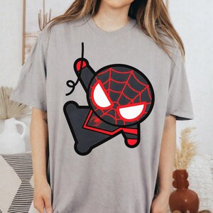 May include: A light grey short-sleeved t-shirt featuring a large, stylised graphic of a cartoon character with a black and red spiderweb pattern, white eyes, and a black spider emblem on the chest, depicted in a dynamic pose with a web line.