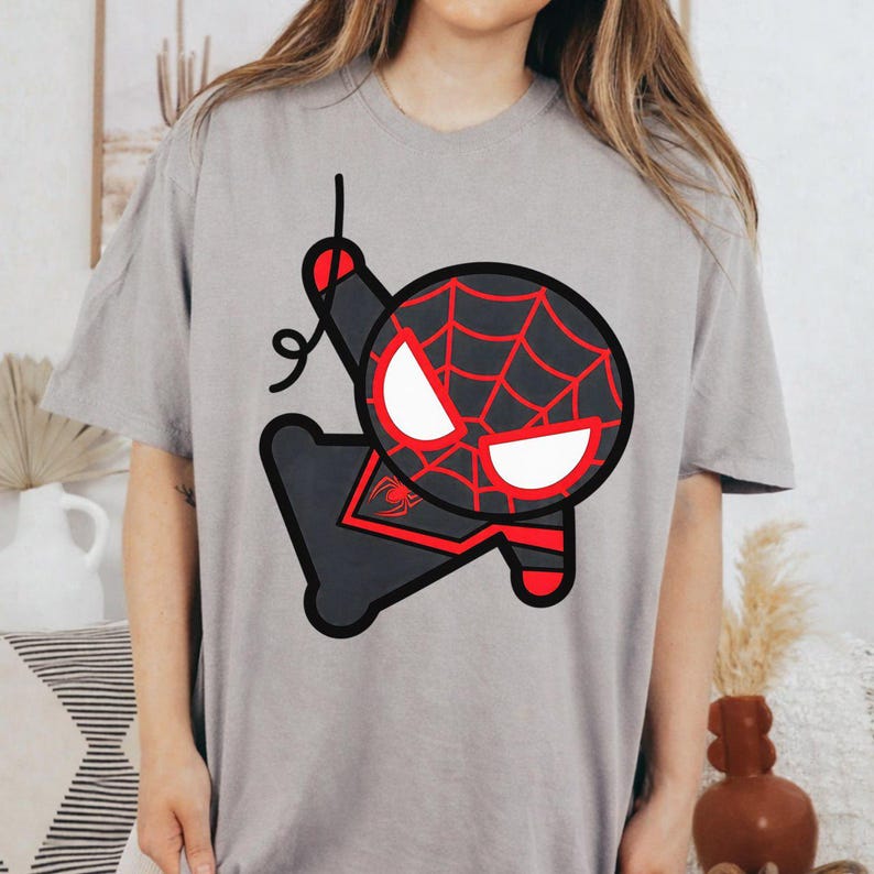 May include: A light grey short-sleeved t-shirt featuring a large, stylised graphic of a cartoon character with a black and red spiderweb pattern, white eyes, and a black spider emblem on the chest, depicted in a dynamic pose with a web line.