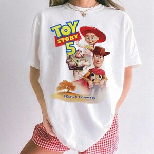 May include: A white t-shirt featuring a colorful graphic of Toy Story 5 with characters Woody, Buzz Lightyear, and Jessie. The design includes the text Toy Story 5 and the phrase I Knew It, I Knew You, set against a landscape with a tree.