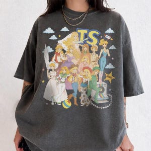 May include: A charcoal gray short-sleeve t-shirt featuring a large graphic print of multiple animated female characters in various outfits. The text TS is printed in large yellow letters at the top, accompanied by clouds, stars, and a rocket.