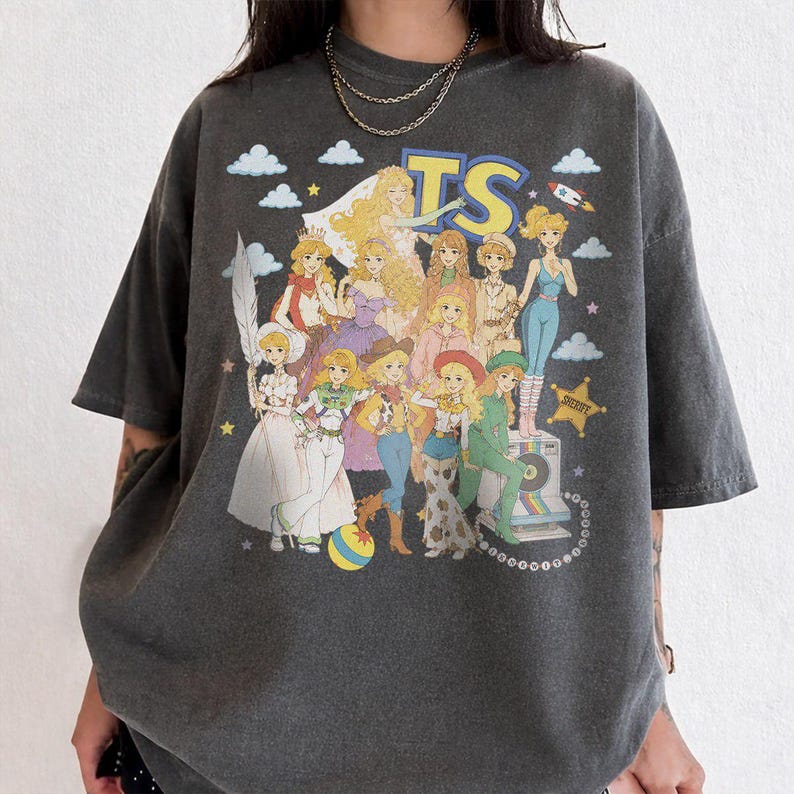 May include: A charcoal gray short-sleeve t-shirt featuring a large graphic print of multiple animated female characters in various outfits. The text TS is printed in large yellow letters at the top, accompanied by clouds, stars, and a rocket.