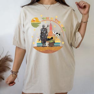Puede incluir: Camiseta beige con un dise&ntilde;o de The Mandalorian y Grogu con globos de Mickey Mouse. El dise&ntilde;o incluye el texto "THIS IS THE WAY" en arco, con un fondo retro de puesta de sol y castillo.