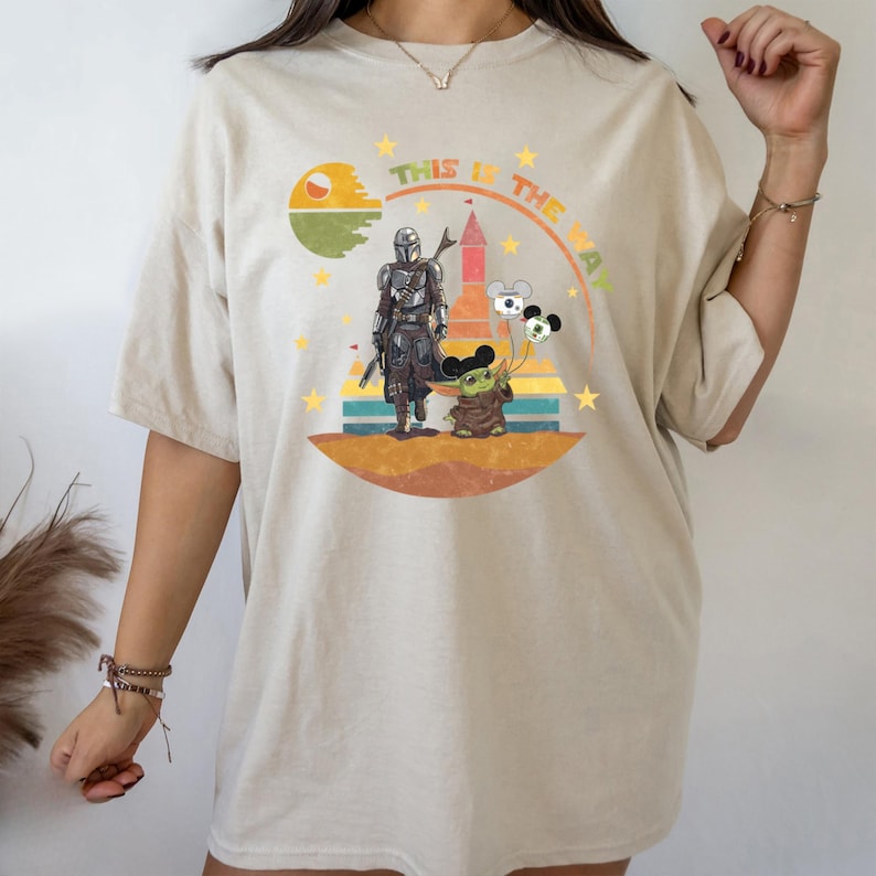 Puede incluir: Camiseta beige con un dise&ntilde;o de The Mandalorian y Grogu con globos de Mickey Mouse. El dise&ntilde;o incluye el texto "THIS IS THE WAY" en arco, con un fondo retro de puesta de sol y castillo.