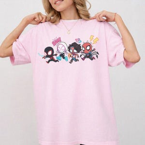 May include: A light pink short-sleeved t-shirt featuring a graphic print of four stylized cartoon characters in action poses. The design includes the text BAM! and question marks, with characters in black, white, red, and blue color schemes.