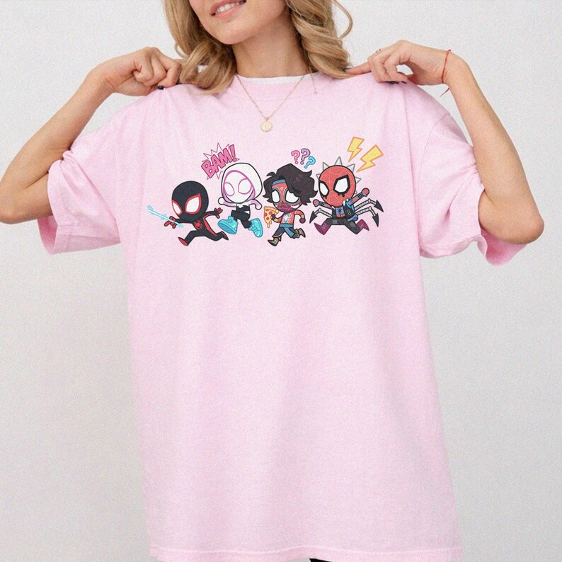 May include: A light pink short-sleeved t-shirt featuring a graphic print of four stylized cartoon characters in action poses. The design includes the text BAM! and question marks, with characters in black, white, red, and blue color schemes.