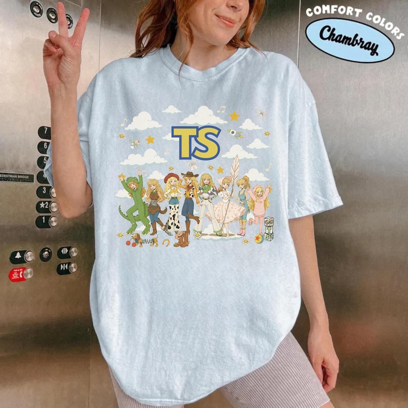 May include: A light blue short-sleeve t-shirt featuring a graphic print of multiple anime-style characters in various costumes against a sky background with clouds and stars. The letters TS are printed in large yellow font above the characters.