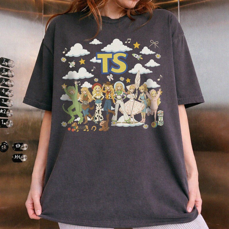 May include: A charcoal gray short-sleeve t-shirt featuring a graphic print of multiple cartoon characters in various outfits. The text TS is printed in large yellow letters at the top, surrounded by white clouds, stars, musical notes, and a rocket.